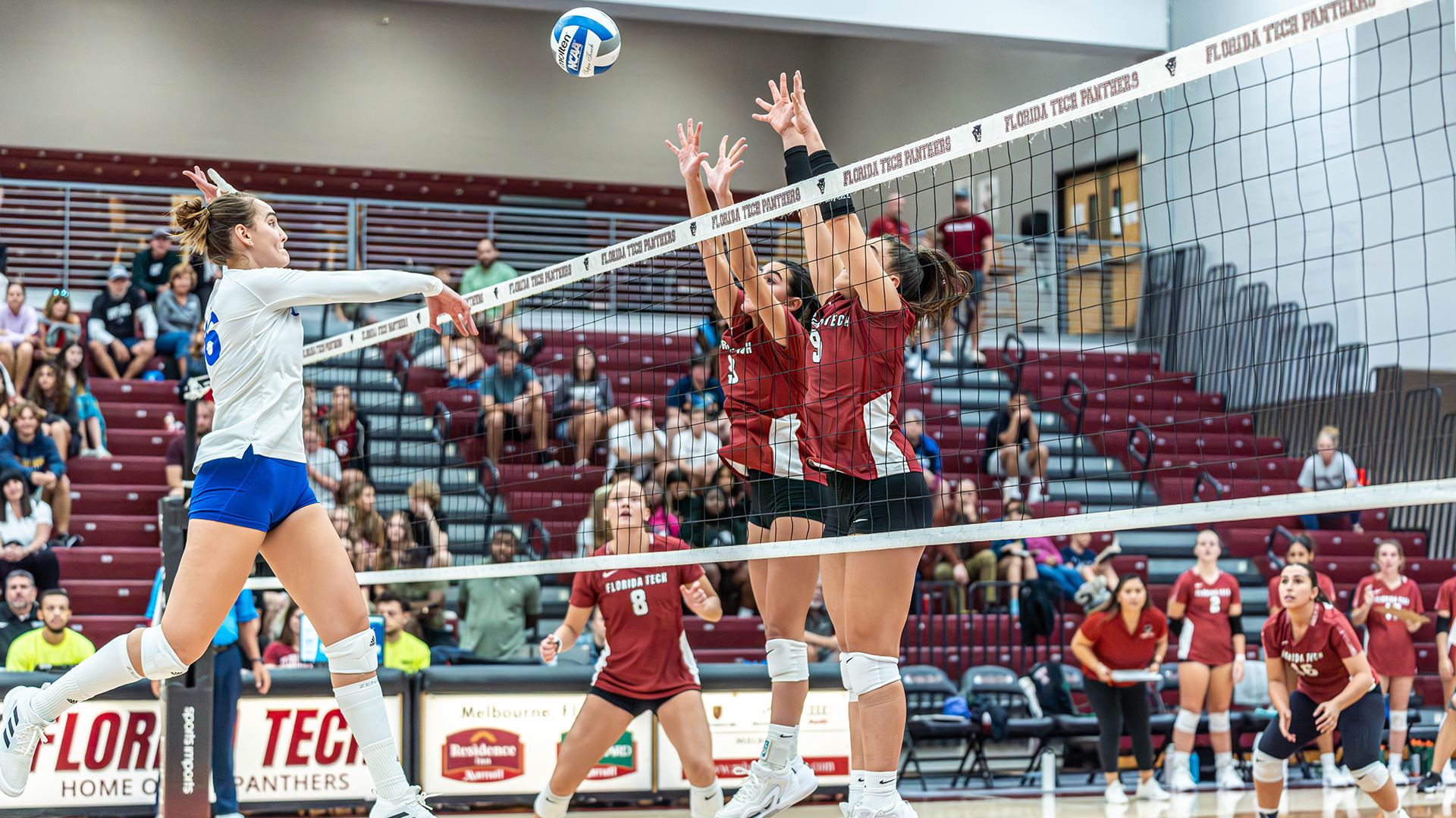 A volleyball match between Florida Tech and another team. A player in a white jersey is spiking the ball over the net while two Florida Tech players in red jerseys are attempting to block. The background shows spectators and other players watching the game